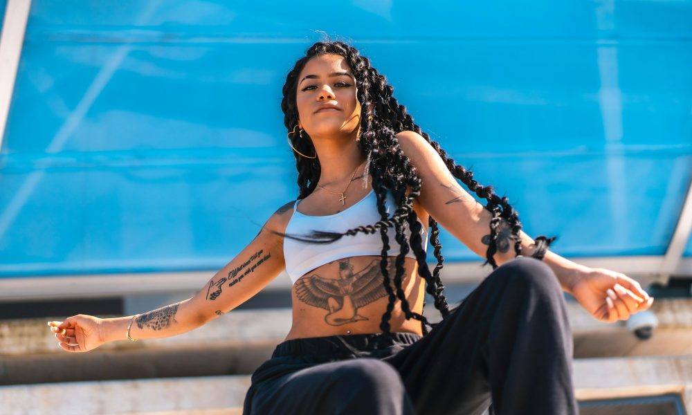 Young Hispanic woman with long braids and tattoos posing under the sunlight